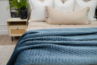 A bed made with tan zipper bedding and an assortment of neutral throw pillows and a blue velvet blanket.
