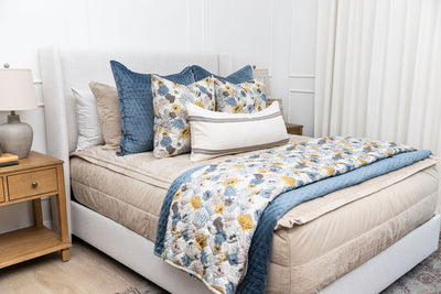 A brightly lit bedroom with a bed made with tan zipper bedding and an assortment of neutral floral and blue velvet throw pillows and blankets.