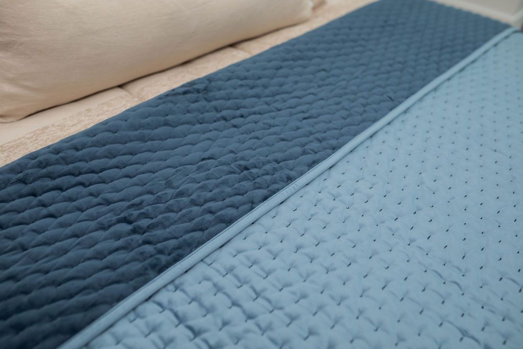 A bed made with tan zipper bedding a blue velvet blanket folded over to reveal the other side.