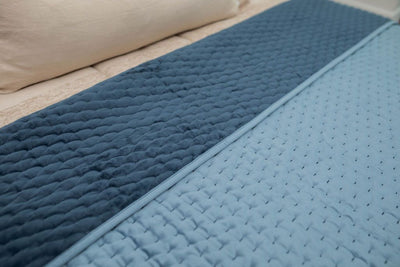 A bed made with tan zipper bedding a blue velvet blanket folded over to reveal the other side.