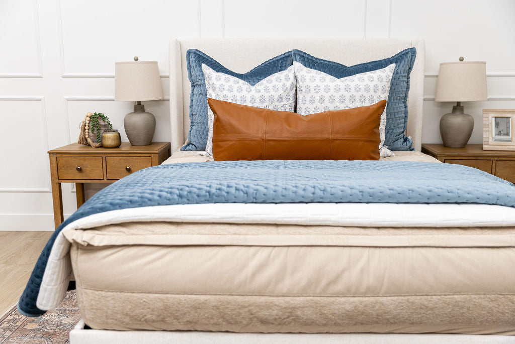 A bed made with tan faded stripes zipper bedding and an assortment of neutral and blue velvet throw pillows and blankets.