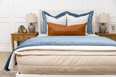 A bed made with tan faded stripes zipper bedding and an assortment of neutral and blue velvet throw pillows and blankets.