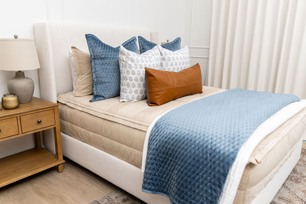 A brightly lit bedroom with a bed made with tan faded stripes zipper bedding and an assortment of neutral and blue velvet throw pillows and blankets.