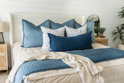 A bed made with tan faded stripes zipper bedding and an assortment of neutral and blue velvet pattern throw pillows and matching blankets.