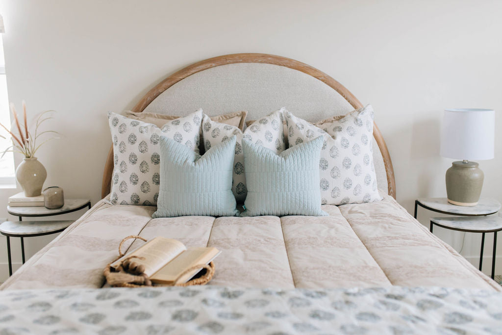 A brightly lit bedroom with a bed made with tan faded stripes zipper bedding and an assortment of neutral and light blue and gray pattern throw pillows and blanket.