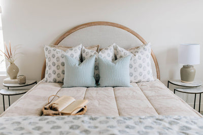 A brightly lit bedroom with a bed made with tan faded stripes zipper bedding and an assortment of neutral and light blue and gray pattern throw pillows and blanket.