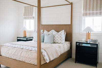 A brightly lit bedroom with a bed made with tan faded stripes zipper bedding and an assortment of neutral and gray floral pattern pillows and blankets.