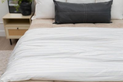 A close up of a bed made with tan zipper bedding and an assortment of neutral pillows and a white and light blue striped blanket.