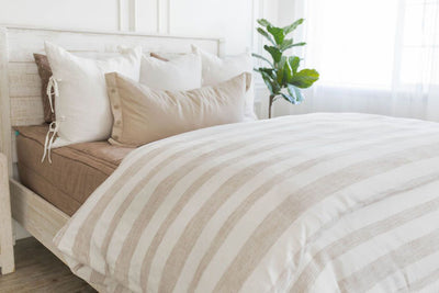 A brightly lit bedroom with a bed made with brown zipper bedding and a woven ivory and tan striped zipper duvet folded half way down the bed and adorned with neutral pillows.