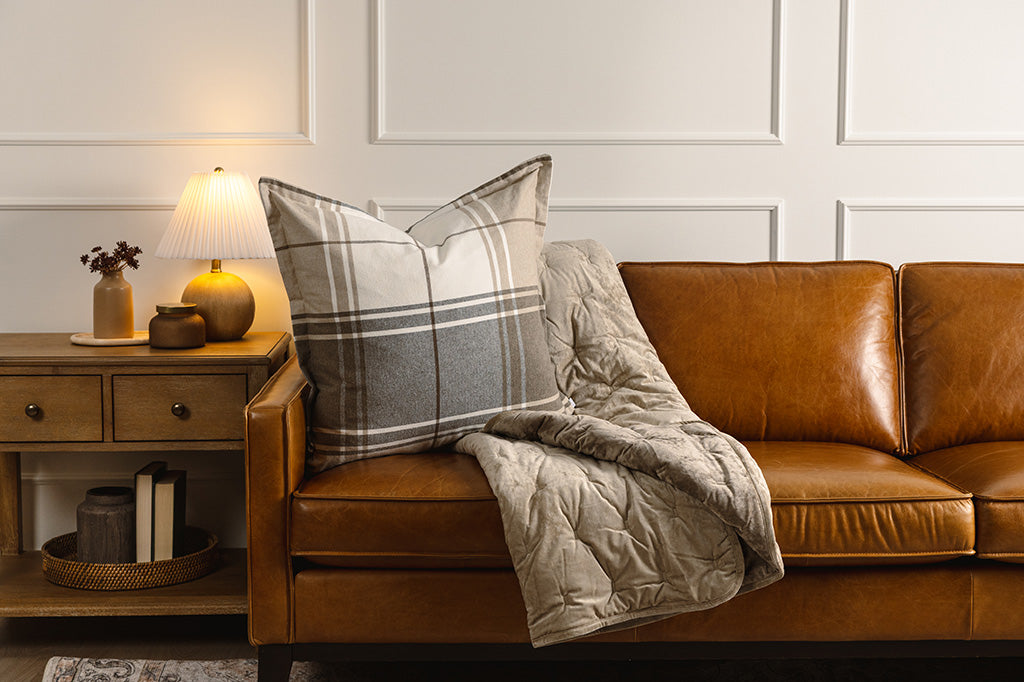 A brown, gray, and white plaid pillow and a brown velvet blanket on a leather couch.