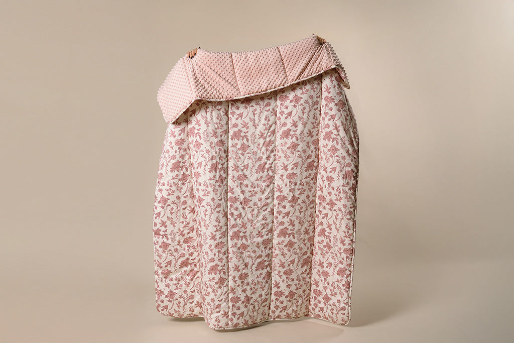 A white and pink, floral and minky blanket being held up by a women on a cream backdrop. 