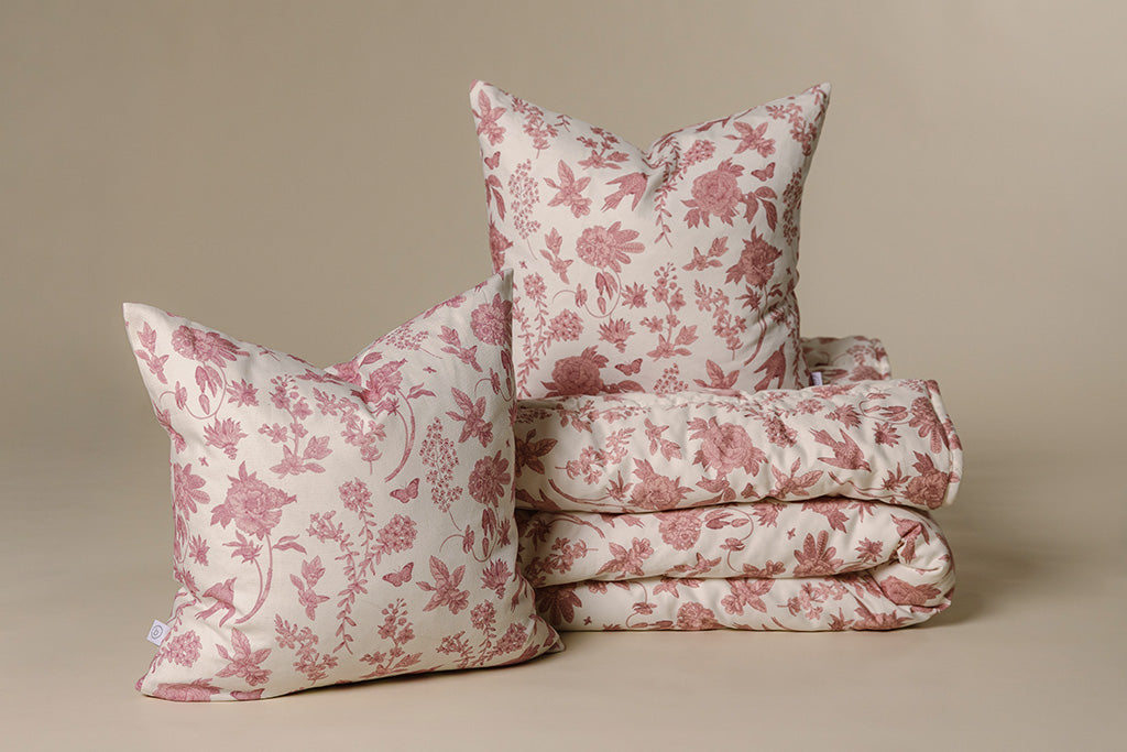 White and pink floral blanket and pillows on a cream paper backdrop.