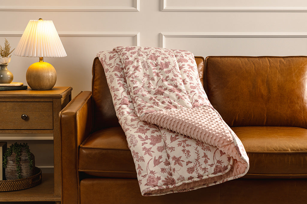 A white pink, floral and minky blanket on an orange leather couch, next to a night stand with a lamp. 