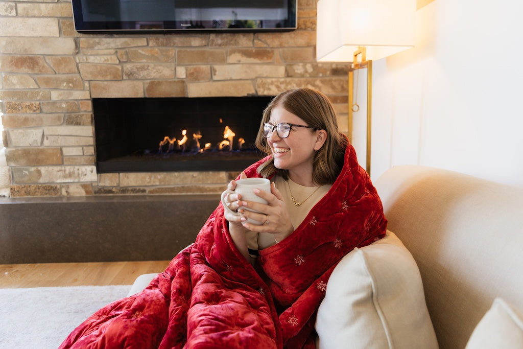 Woman cuddled up in a red and white snowflake blanket by the fireplace. 