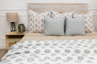 A bed made with tan zipper bedding and an assortment of neutral and gray floral pattern throw pillows and blanket.