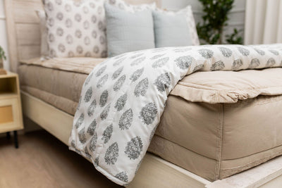 A side view of a bed made with tan zipper bedding with a gray floral pattern white blanket folded over the end of the bed.
