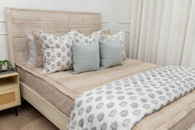 A side view of a bed made with tan zipper bedding and an assortment of neutral and gray floral pattern throw pillows and blanket.