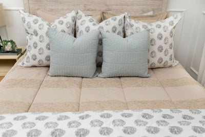 A close up view of a bed made with tan striped zipper bedding and an assortment of light blue and gray floral pattern pillows and blanket.