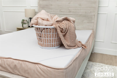 A bed made with just the fitted bedding base around the sides of the mattress, with the top comforter and ZipBase sheet in a basket on top of the bed.