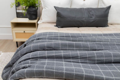A close up of a bed made with tan zipper bedding and neutral pillows with a deep gray and white grid stitching pattern blanket at the end of the bed. 