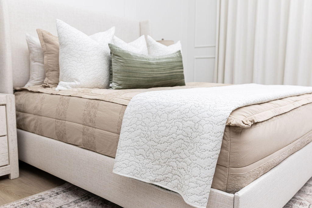 Side view of a bed made with tan zipper bedding with white stitched throw pillows, a green lumbar pillow, and a white stitched throw blanket.