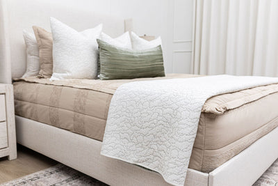 Side view of a bed made with tan zipper bedding with white stitched throw pillows, a green lumbar pillow, and a white stitched throw blanket.