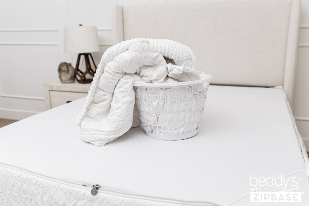 A bed made with just the fitted bedding base around the sides of the mattress, with the top comforter and ZipBase sheet in a basket on top of the bed.