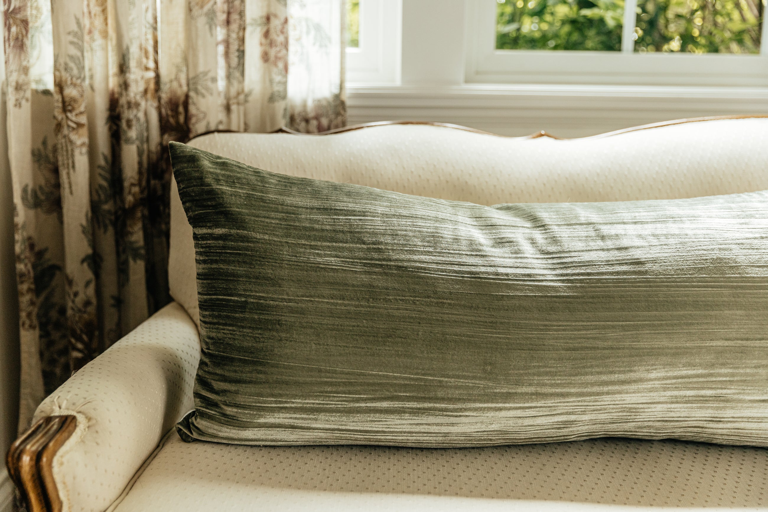 A green XL velvet lumbar pillow arranged on a cream antique couch with a window and curtains behind it.