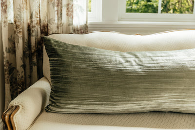 A green XL velvet lumbar pillow arranged on a cream antique couch with a window and curtains behind it.