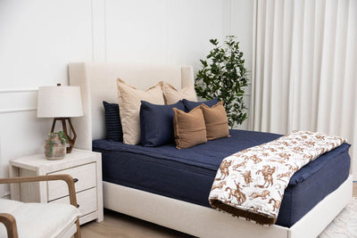 A brightly lit bedroom with a bed made with denim blue zipper bedding and an assortment of neutral pillows and cowboy western printed blanket.