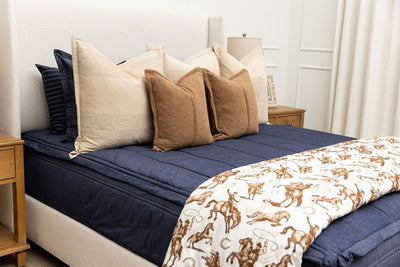 A brightly lit bedroom with a bed made with dark denim blue zipper bedding and an assortment of neutral pillows and a cowboy western printed blanket.