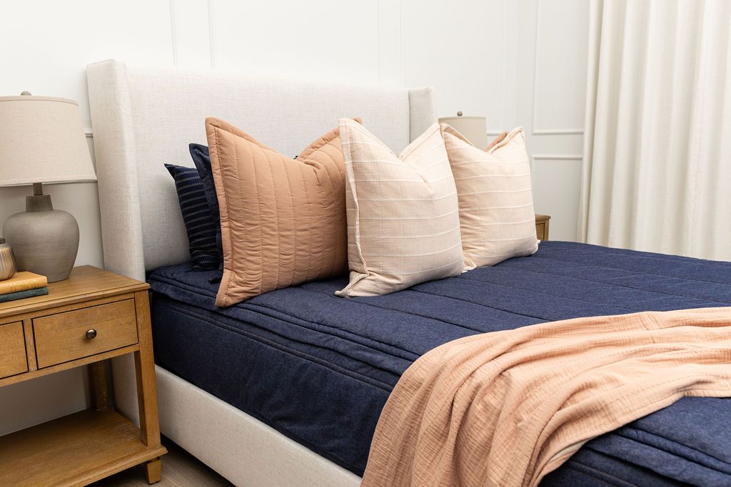 A brightly lit bedroom with a bed made with dark denim blue zipper bedding and an assortment of neutral and terracotta colored pillows and blanket.