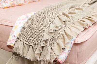 A side view of a bed made with pink zipper bedding with a tan waffle textured throw blanket folded over the end of the bed on top of a multicolored floral blanket.