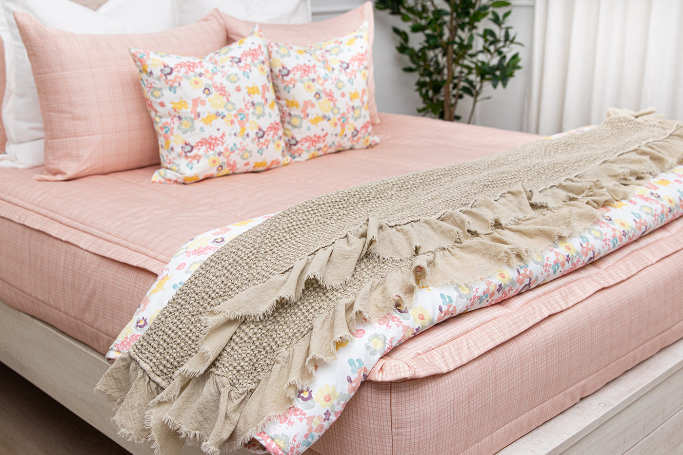A bed made with pink zipper bedding and multicolored floral pillows and blanket with a tan waffle textured throw blanket at the end of the bed.