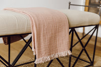 A clay/pink colored throw blanket folded and draped over an upholstered bench.