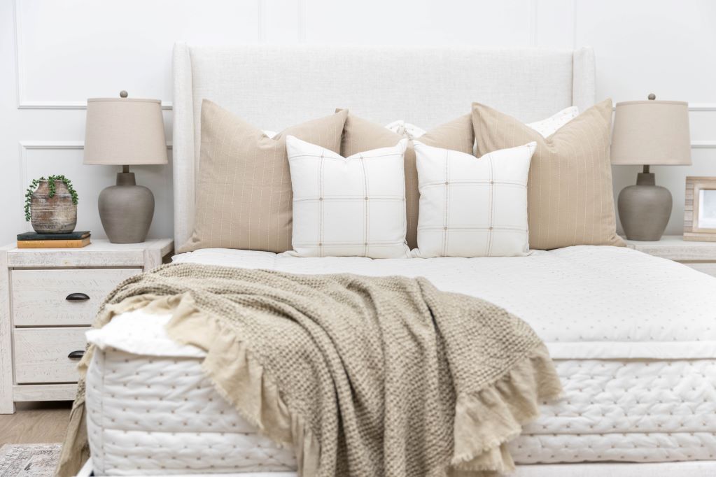 A lightly lit bedroom with a bed made with cream embroidered zipper bedding, decorated with neutral throw pillows and blankets.