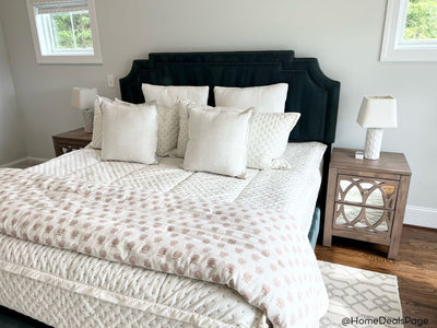 A queen bed made with cream embroidered zipper bedding and adorned with neutral pattern pillows and a light floral pink blanket.
