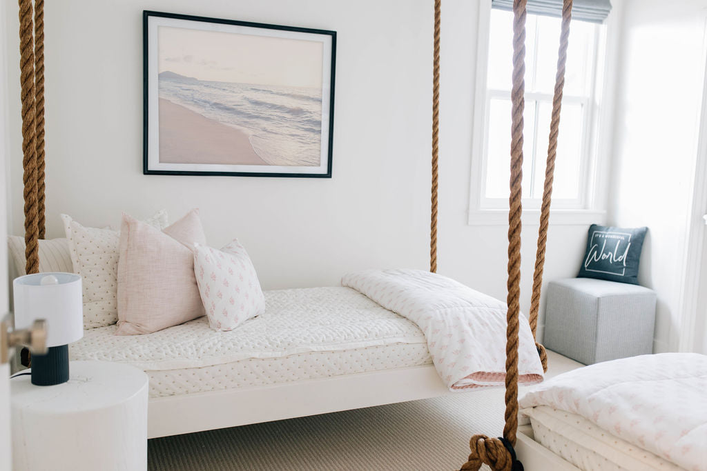 A twin hanging bed made with cream embroidered zipper bedding and adorned with light pink pattern pillows and blanket.
