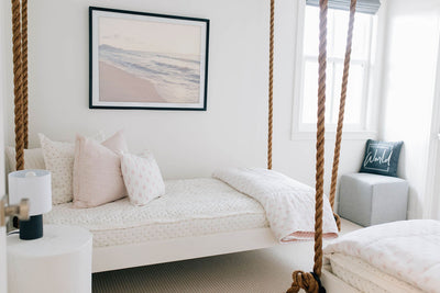 A twin hanging bed made with cream embroidered zipper bedding and adorned with light pink pattern pillows and blanket.