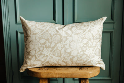 A floral lumbar pillow on a wood stool in front of a green wall.