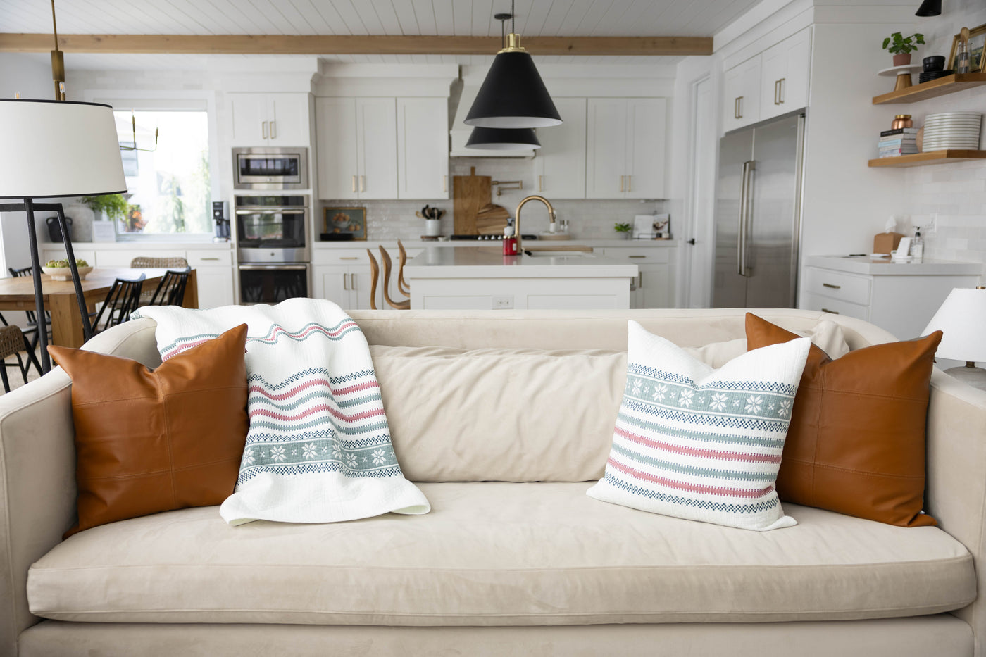 A cream couch in a living room with leather brown and holiday pattern white striped pillows and matching blanket.