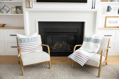 Two living room chairs by a fireplace with a holiday white striped pillow and matching blanket on them.