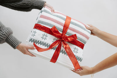 A red, green, and snowflake striped pattern white holiday blanket folded and wrapped in a red ribbon with two people holding it.