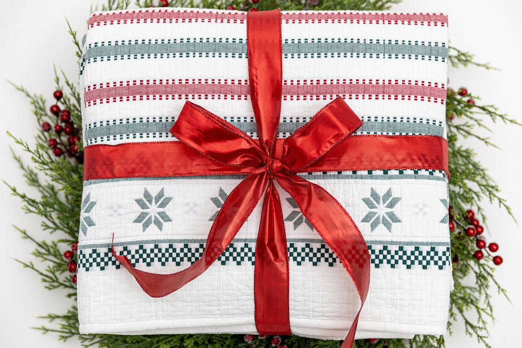 A red, green, and snowflake striped pattern white holiday blanket folded and wrapped in a red ribbon on top of a green wreath.