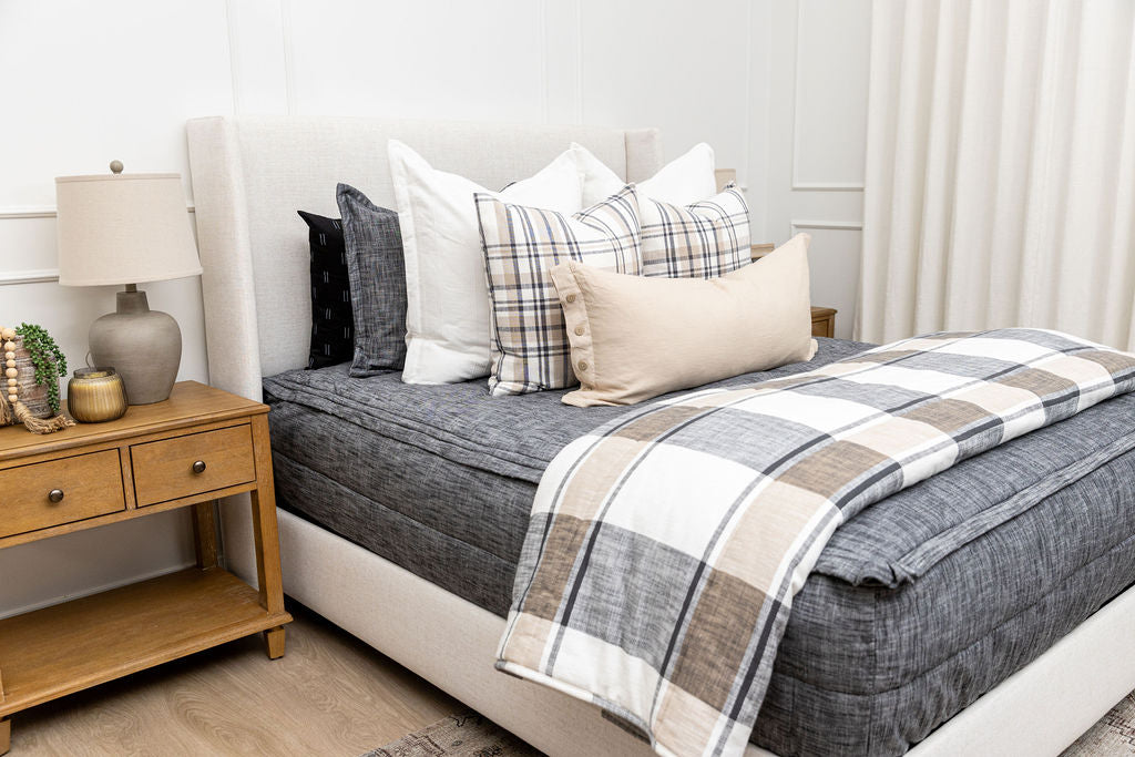 A queen bed made with dark charcoal gray zipper bedding and an assortment of plaid and neutral pillows and blanket.