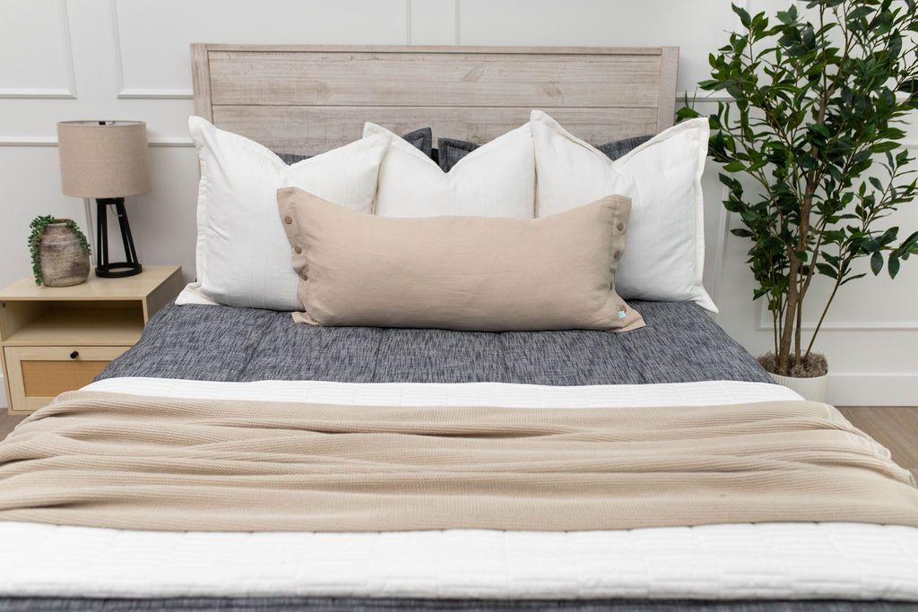 A close up view of a bed made with dark charcoal gray zipper bedding and neutral tan and white throw pillows and blankets.