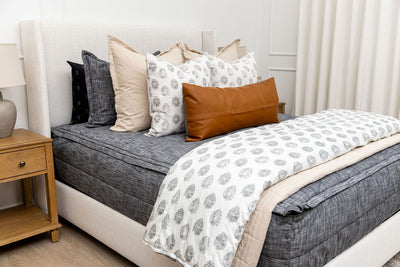 A queen bed made with dark charcoal gray zipper bedding and an assortment of neutral tan and gray pattern pillows and blankets.