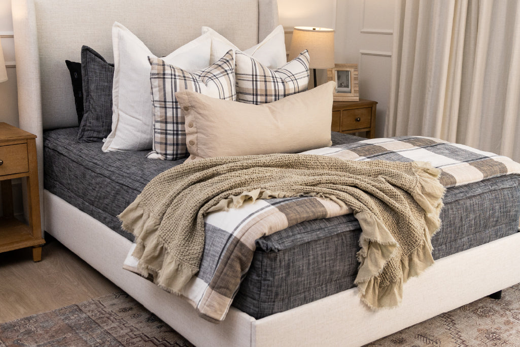 A bed made with dark charcoal gray zipper bedding and an assortment of neutral and plaid throw pillows and blanket.