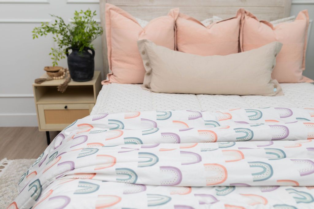 A close up of a bedding made with cream embroidered zipper bedding with peach and neutral pillows and a pastel rainbow pattern blanket at the end of the bed.