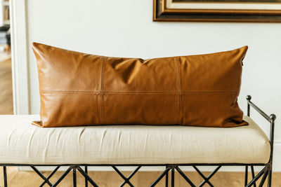 A neutral leather XL lumbar pillow with stitching arranged on a cream bench.